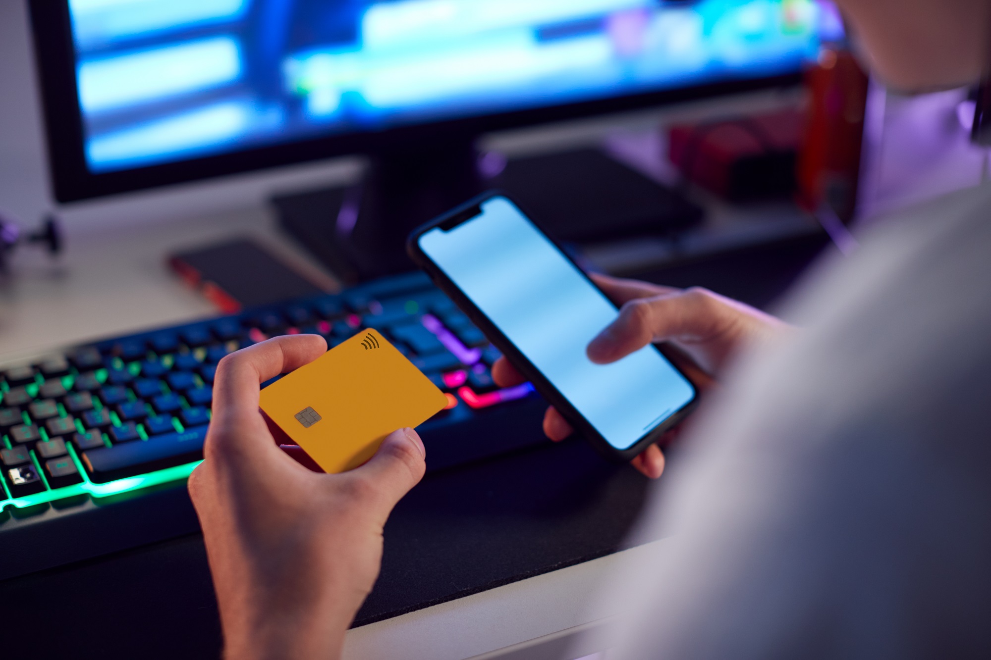 Imagem close-up nas mãos de um homem, na direita ele segura um cartão de crédito, enquanto na esquerda segura um celular. Ao fundo está um teclado e um monitor.