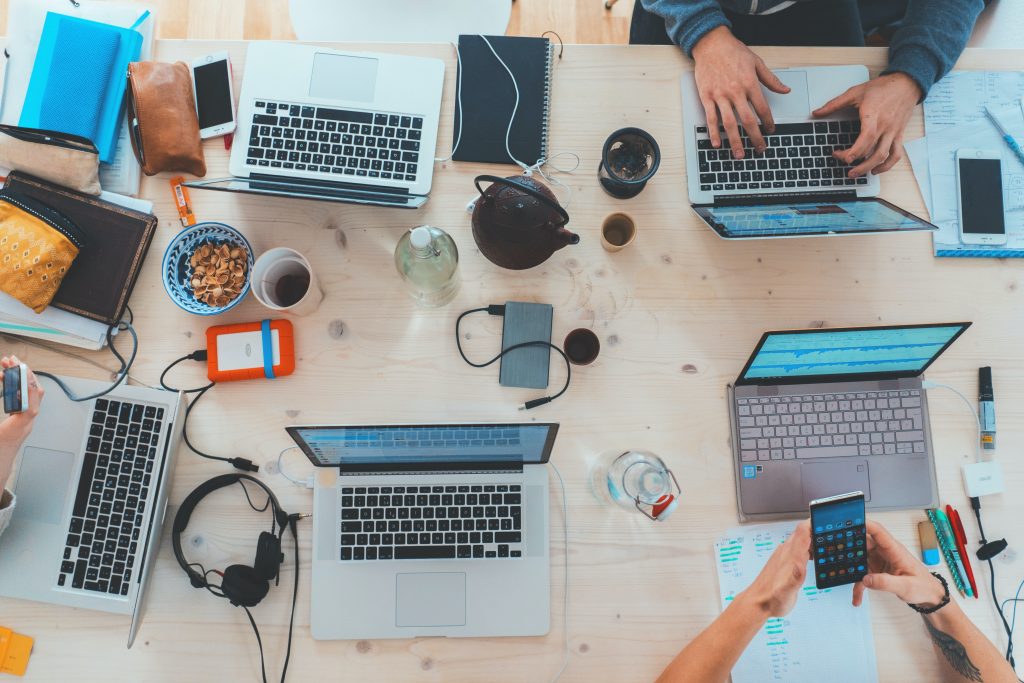 Foto vista aérea, com diversos dispositivos por cima, um ao lado do outro, entre eles smartphones e notebooks em cima de uma mesa, dois deles sendo mexidos por duas pessoas