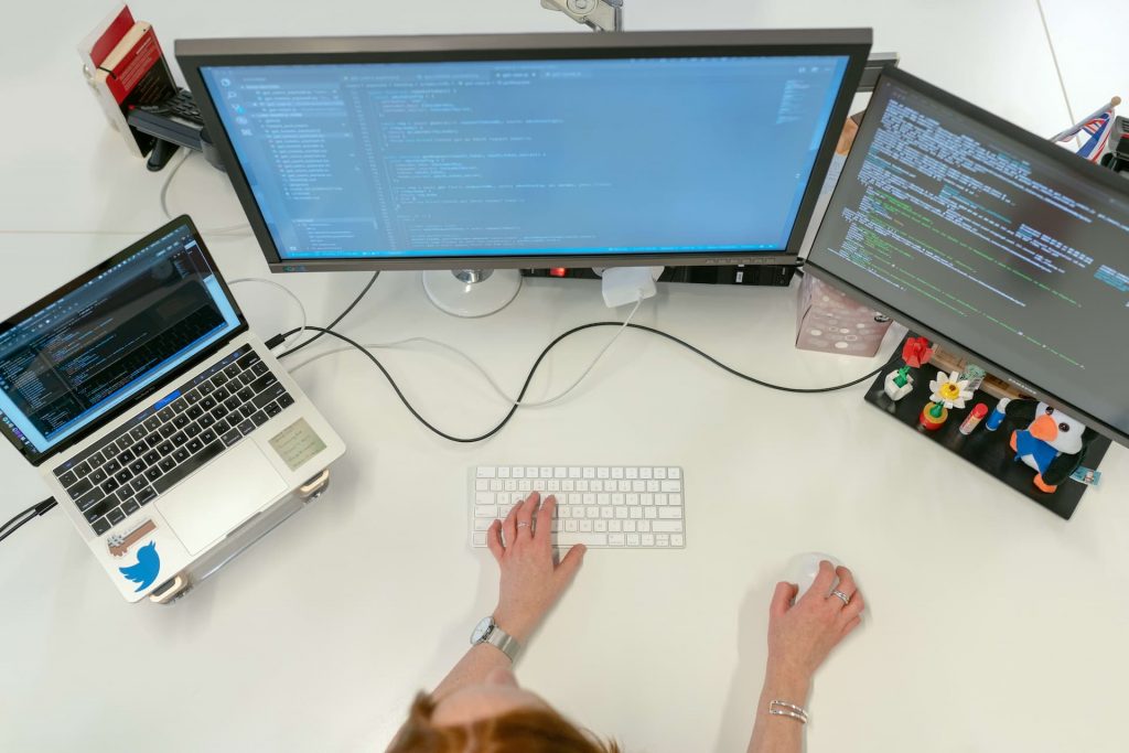 Imagem vista da perspectiva de cima mostra uma mulher mexendo em um notebook ligado a outras duas telas, programando