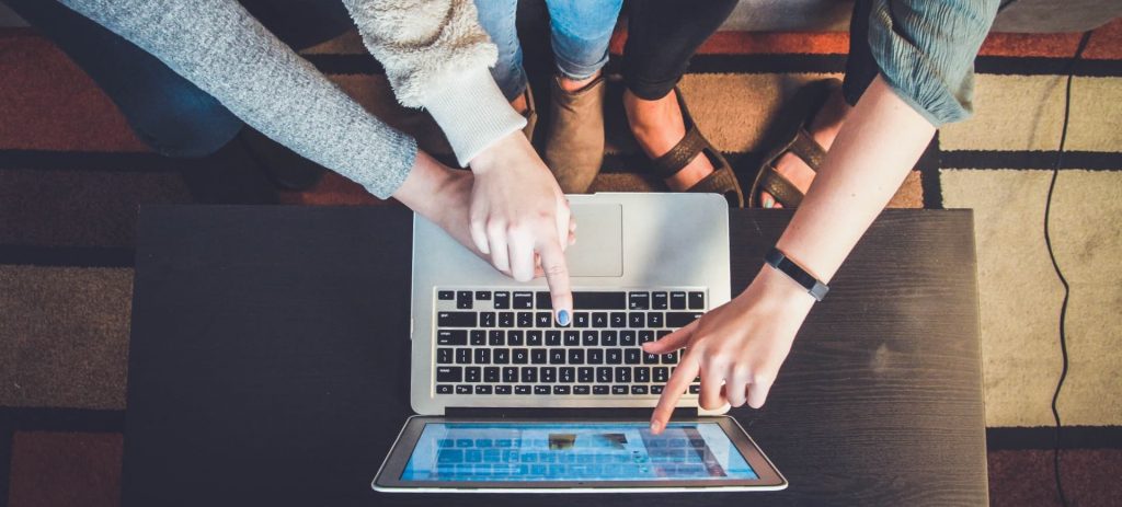 Imagem com vista aérea mostra um notebook em cima de uma mesa e três mãos, de três diferentes jovens, apontando para a tela como se estivessem escolhendo ou discutindo o que fazer com a informação na tela