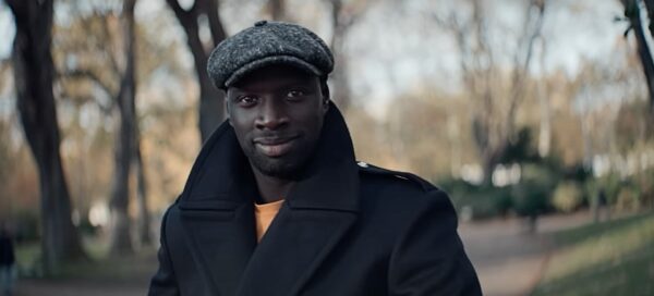 Cena de Lupin, série francesa da Netflix; na imagem o ator principal Omar Sy com uma boina e casaco, ao fundo uma paisagem de árvores