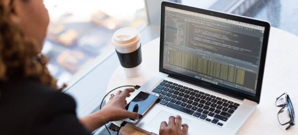 Na foto aparece o rosto de uma mulher de perfil, bem como suas mãos, sendo uma posicionada no mouse pad de um notebook, enquanto a outra está segurando um celular; a tela do notebook exibe código de programação; ela representa as mulheres na tecnologia