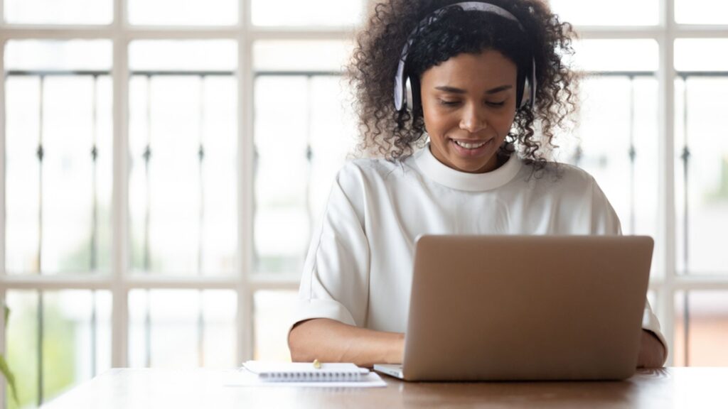 Imagem mostra uma mulher de cabelos cacheados e blusa branca sentada em uma mesa digitando em um notebook, representando mulheres na área de tecnologia