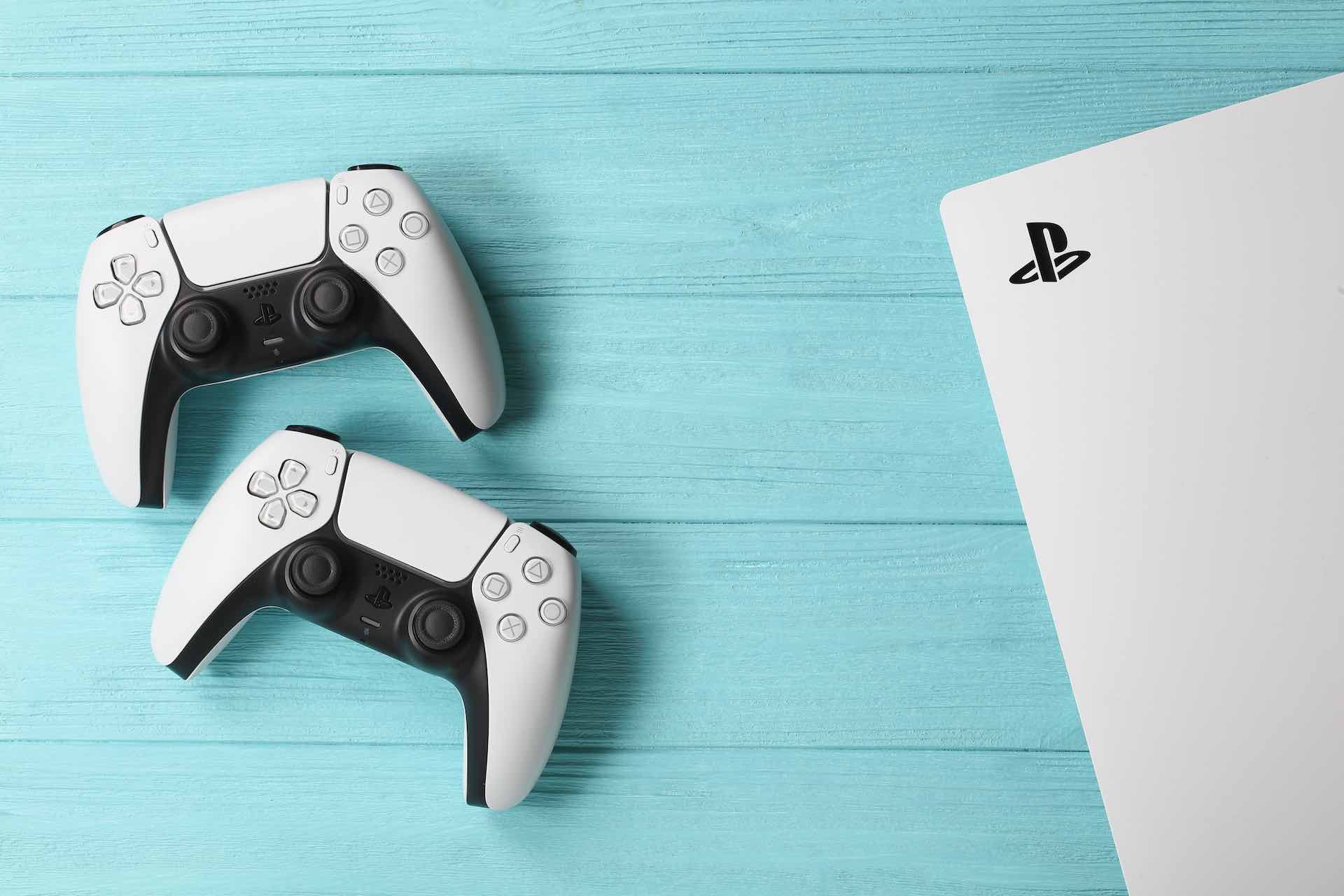 Em um fundo azul, a imagem exibe dois controles brancos do PlayStation 5 ao lado de um pedaço do console da Sony