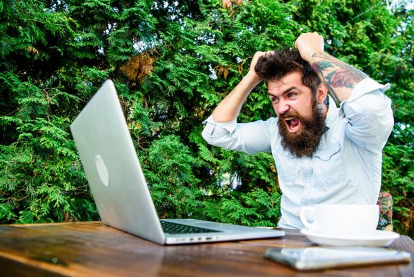 Imagem mostra um homem de barba grande, camisa branca e tatuagem nos braços nervoso com o seu notebook