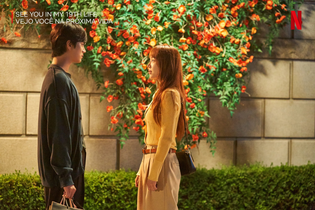 Cena do novo dorama da Netflix chamado Vejo Você na Próxima Vida, com os dois atores principais de perfil, olhando um para o outro, em um cenário com uma parede com flores ao fundo