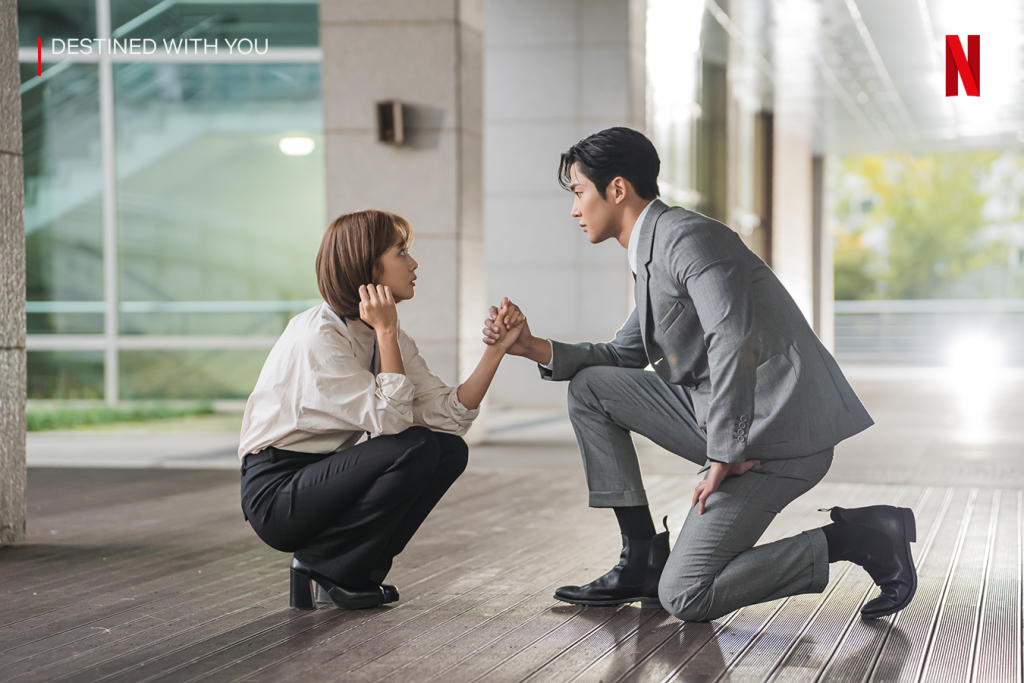 Cena do dorama Destined with You, da Netflix, onde aparecem os dois atores principais agachados no chão de um escritório, vestidos com roupas formais e se olhando, os dois formam o par romântico heterossexual