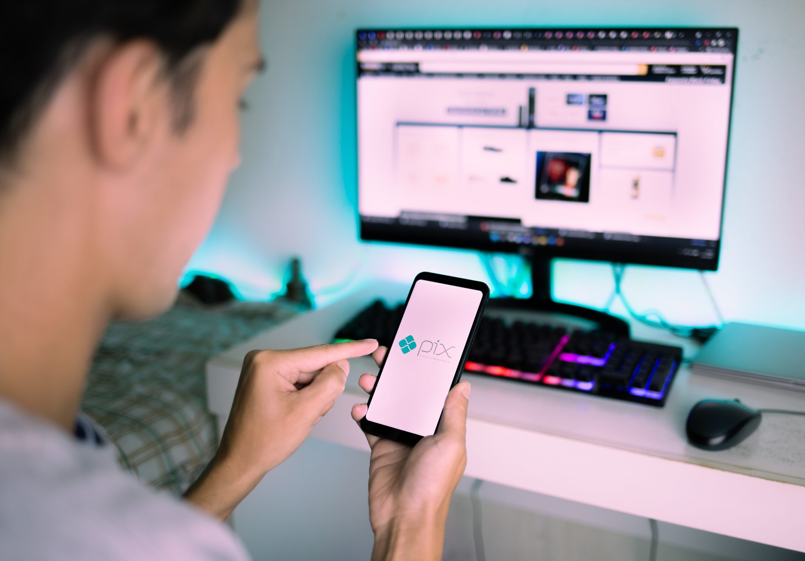 Imagem mostra um jovem usando o PIX no celular à frente de um computador
