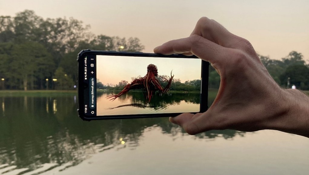 Vista da obra de realidade virtual de Fernando Velázquez, parte da exposição Realidades e Simulacros do Museu de Arte Moderna de São Paulo (MAM). A foto mostra, ao fundo, o lago do Parque Ibirapuera e, à frente, uma mão segurando um smartphone que interage com o lago e mostra uma escultura em realidade aumentada na tela