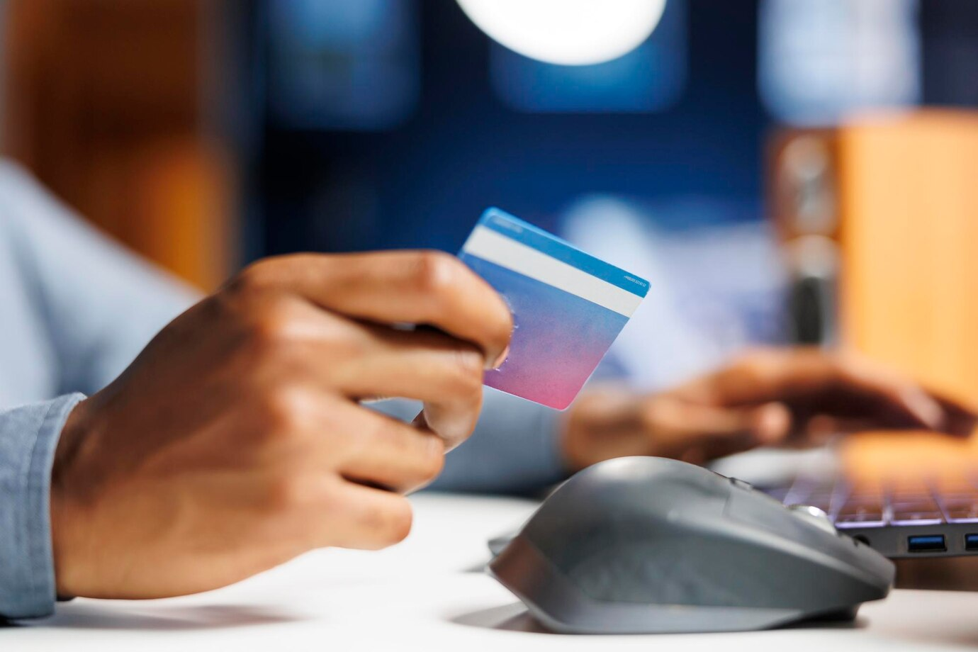 Uma imagem close-up na mão de um homem. Ele está segurando um cartão de crédito enquanto faz compras online. É possível ver um mouse e um teclado ao fundo.