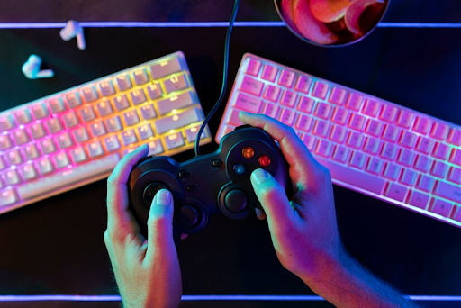 Imagem focada em duas mãos que estão segurando um controle de console. Ao fundo podemos ver dois teclados gamers.