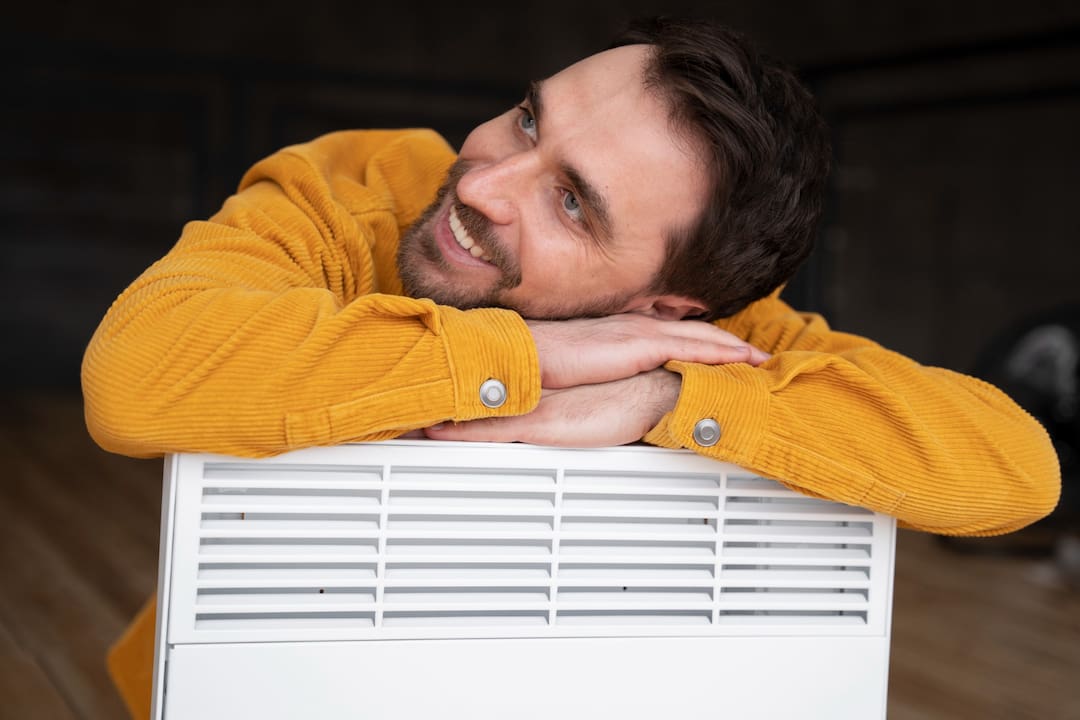 Homem sorridente com barba e cabelo castanho escuro, vestindo um suéter amarelo mostarda, apoiado sobre um aparelho de ar condicionado portátil branco. Ele parece relaxado e contente, com um fundo levemente desfocado.